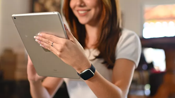 Mulher branca, cabelo castanho escurro, ela está sorrindo com um tablet na mão, em uma das mãos tem um anel cinza, também está com um relógio preto no braço esquerdo