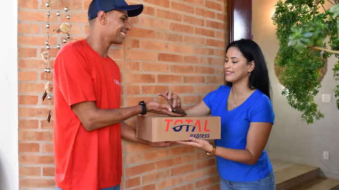 Homem entregando uma caixa pequena com o logotipo da Total Express para uma mulher, o homem é preto e está de camiseta vermelha e boné azul, a mulher é branca e está com camiseta azul 