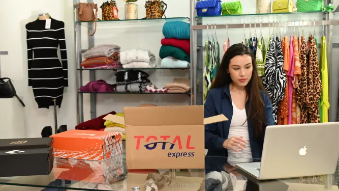 Empresária gerenciando pedidos e logística no computador, com caixas da Total Express, representando a gestão de e-commerce.