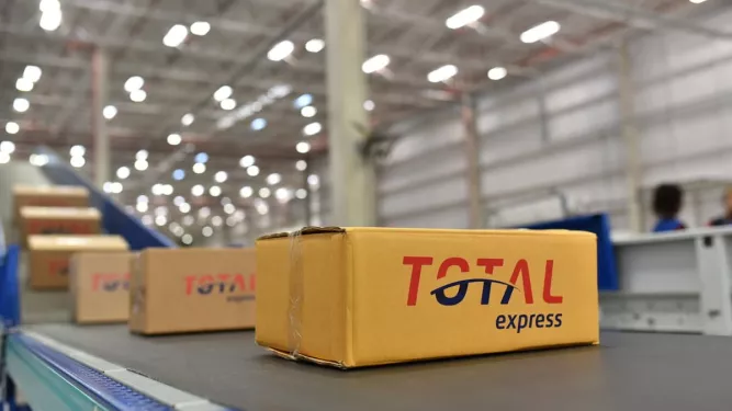 Caixa com o logo da Total Express em uma esteira de distribuição, ilustrando o processo de supply chain em um centro de logística.