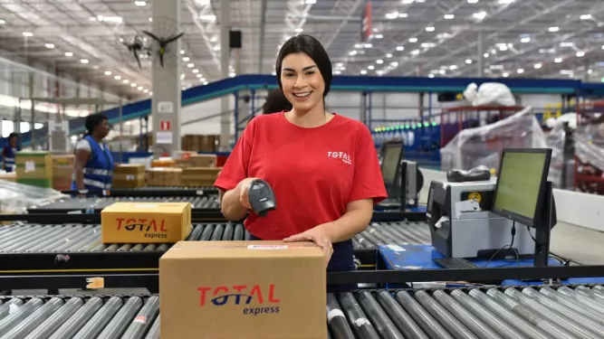 Funcionária da Total Express sorri enquanto escaneia uma caixa em esteira automatizada, ilustrando um ambiente moderno de centro de distribuição e fluxo logístico.
