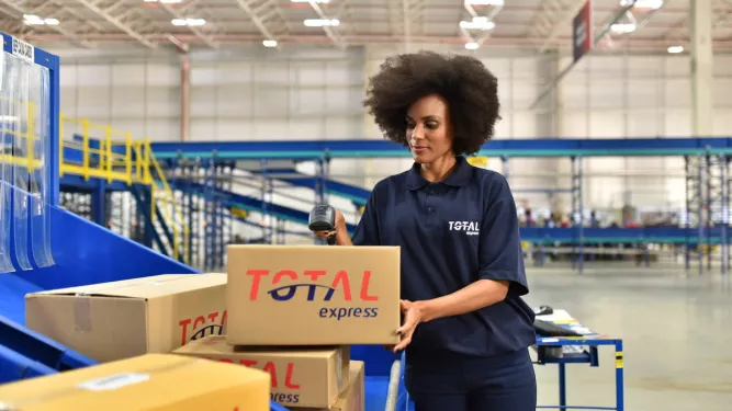 A imagem mostra uma profissional em um armazém manuseando caixas e utilizando um leitor de código de barras, ilustrando o processo logístico relacionado ao frete CIF e FOB.