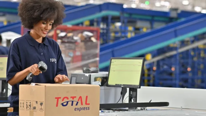 Representação da logística 4.0 com uma operadora fazendo a leitura de uma etiqueta em caixa da Total Express.