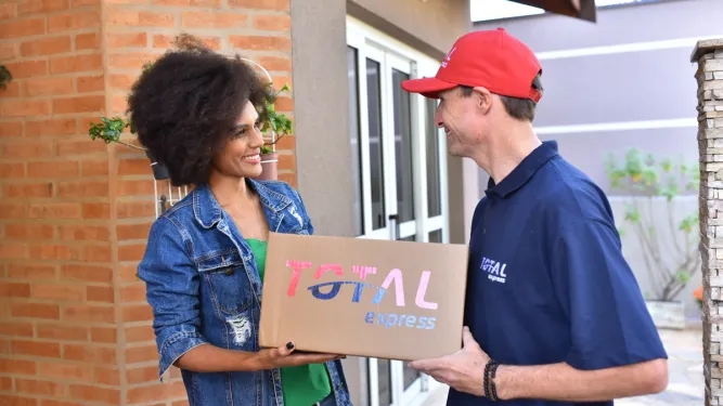 Mulher recebe caixa da Total Express com encomenda. O entregador faz uma logística sustentável