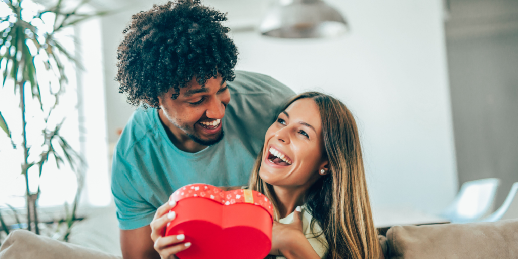 Um casal sorri alegremente enquanto segura uma caixa de presente vermelha em formato de coração. O homem está atrás de uma mulher que segura a caixa, e eles parecem estar em uma sala moderna e iluminada. Uma planta e uma luminária são visíveis ao fundo.