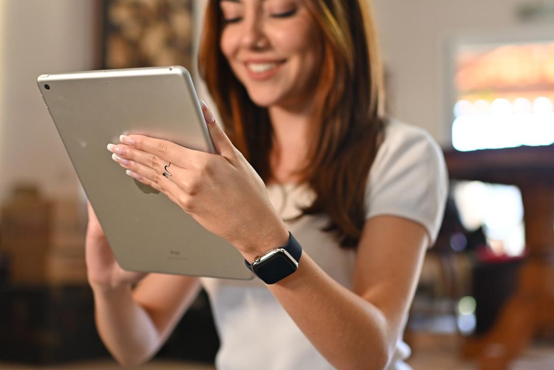 Mulher branca, cabelo castanho escurro, ela está sorrindo com um tablet na mão, em uma das mãos tem um anel cinza, também está com um relógio preto no braço esquerdo
