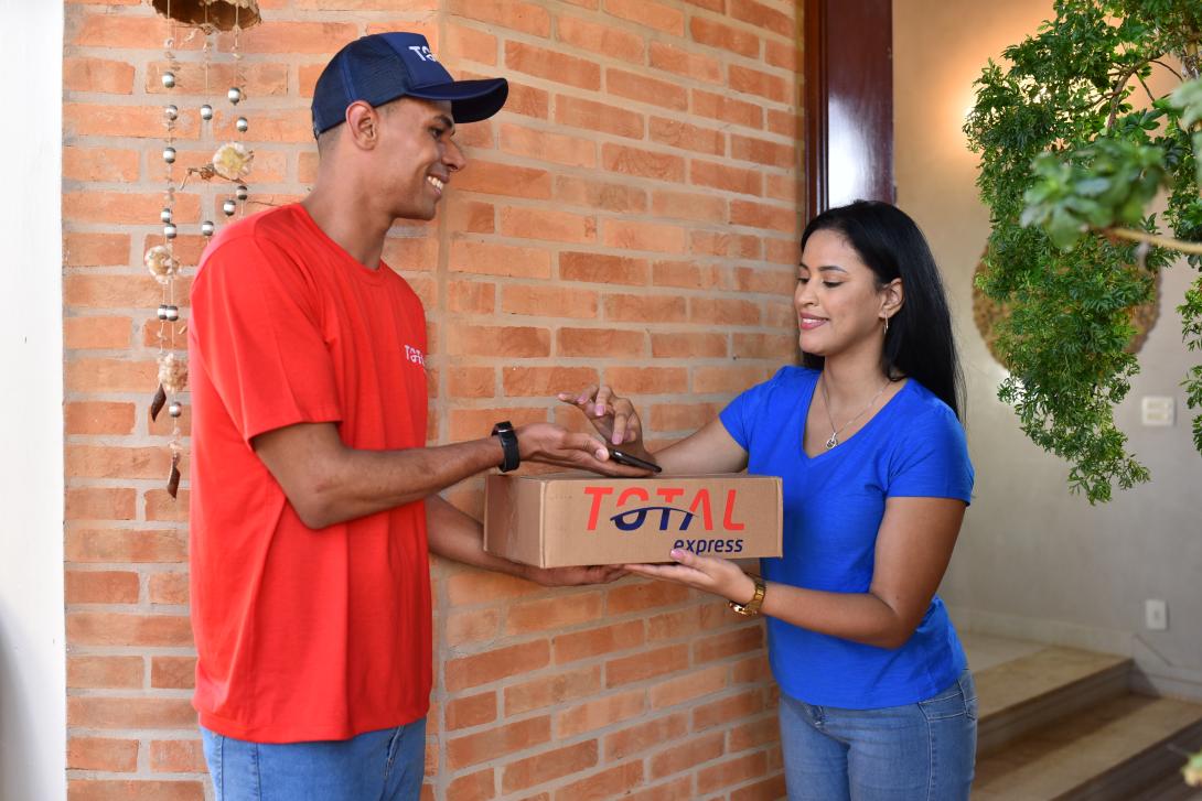 Homem entregando uma caixa pequena com o logotipo da Total Express para uma mulher, o homem é preto e está de camiseta vermelha e boné azul, a mulher é branca e está com camiseta azul 