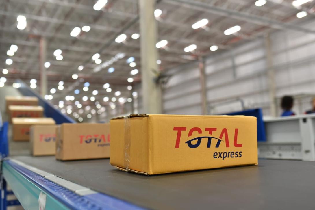 Caixa com o logo da Total Express em uma esteira de distribuição, ilustrando o processo de supply chain em um centro de logística.