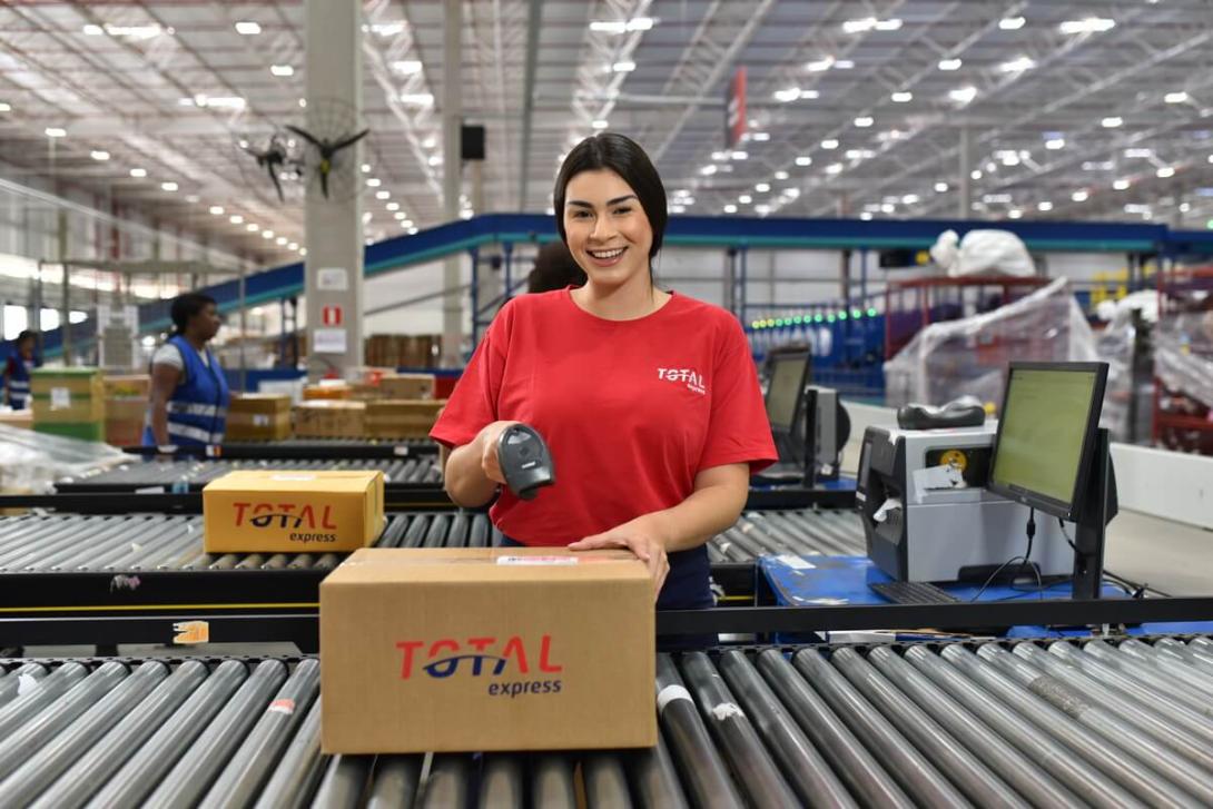 Funcionária da Total Express sorri enquanto escaneia uma caixa em esteira automatizada, ilustrando um ambiente moderno de centro de distribuição e fluxo logístico.
