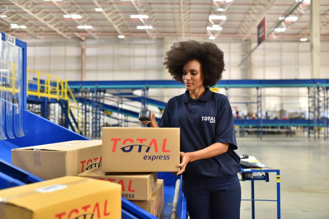 A imagem mostra uma profissional em um armazém manuseando caixas e utilizando um leitor de código de barras, ilustrando o processo logístico relacionado ao frete CIF e FOB.