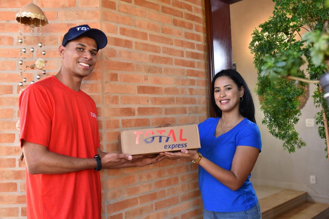 Total Solutions: Entregador da Total Express realizando a entrega de um pacote para o cliente, com ambos sorrindo na porta da residência.