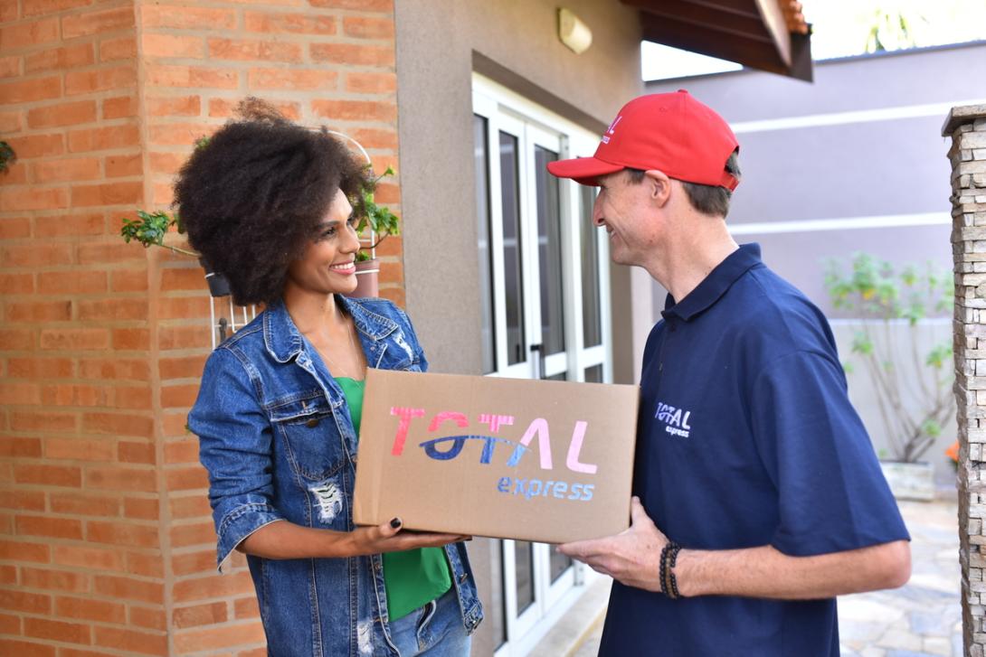 Mulher recebe caixa da Total Express com encomenda. O entregador faz uma logística sustentável